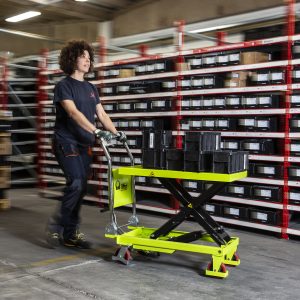 Warehouse worker pushing a bright yellow pallet jack along tall red-shelved racks filled with black bins and boxes.