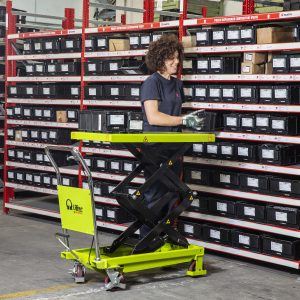 Warehouse worker on a yellow powered pallet lift handling labeled plastic crates on tall red shelving racks.